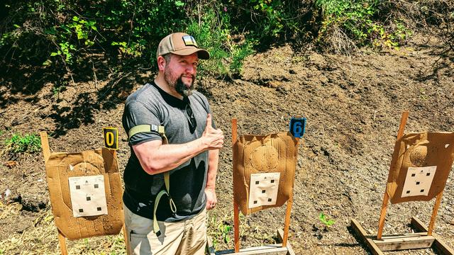 Me in front of a target berm with a USGI sling on my arm, giving a thumbs up, after shooting a string of rounds