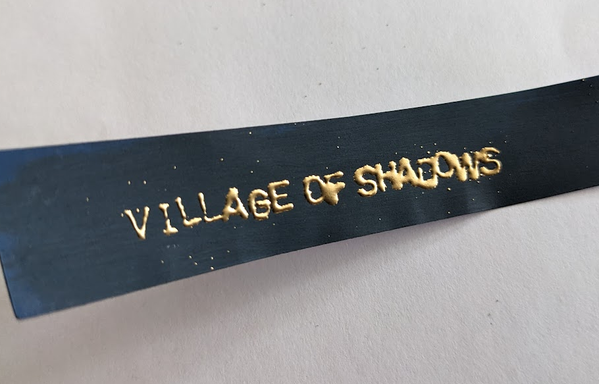Strip of dark blue paper with gold lettering saying 'Village of shadows' in all caps.