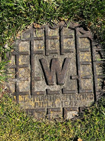 A manhole cover, as they used to be called, a cover for a underground utilities. This is round, surrounded by grass. There is an interesting pattern of lines that creates almost a waffle effect – mostly continuous vertical bars, from which horizontal bars extend, but the horizontals don't quite connect with the next vertical, so it's a broken pattern. In the center is a large sans serif W, presumably representing the name of the foundry, but I haven't figured out what foundry it would have been. Below the W in much smaller letters are two lines of type. The first is "CONTRACTORSFDRYINC" where I think they have abbreviated "foundry" as "FDRY," not so unusual – so it should mean Contractors Foundry, Incorporated. Beneath that, Phila (for Philadelphia), 1942.
