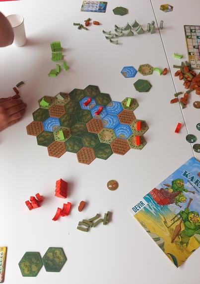 Kakekare tabletop boardgame with some wood miniature artfacts and ressource tokesn, hexagonal.tiles and the rulesheet displaying a maori warrior like kiwi bord in front cover.