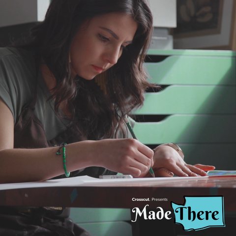 Christie Tirado, a Mexican-American woman with brown hair, wearing a sage green shirt, sits at a table with a pencil, drawing. There is a light green flat-file cabinet behind her.