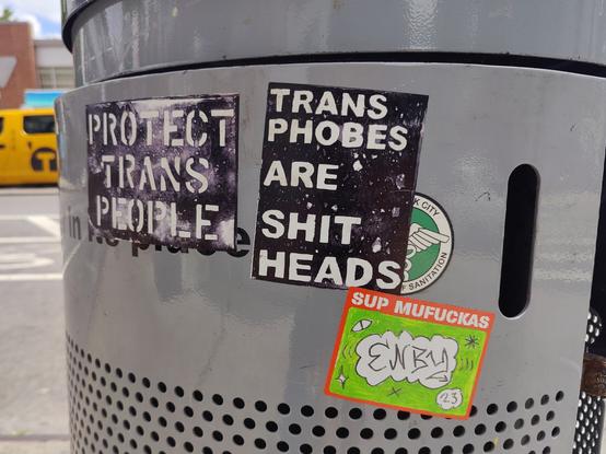A garbage can in Brooklyn with several stickers on them, two are by me and say "protect trans people" and "transphobes are shitheads". The third is by a mysterious artist whose name is presumably Enby and says "sup muhfuckas, enby, 2.3"