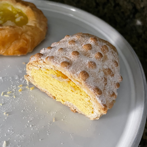 Half of a pan dulce (Mexican sweet bread) with a sugary brown crust covered in large round bumps vaguely resembling the corn kernels of an elote (corn cob). The soft inside of the pastry is a vibrant yellow.