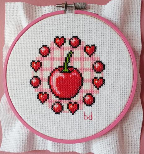 Cross stitch piece of a large red cherry surrounded by red hearts and balls on a pink gingham background, framed in a pink hoop