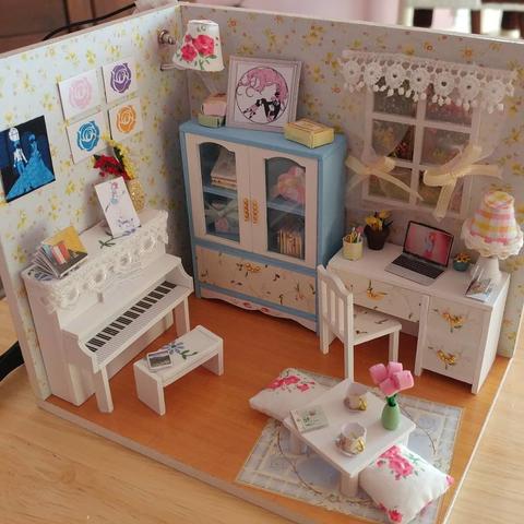 A tiny dollhouse room featuring a white piano, blue and white hutch, white desk, and small white table with pillows to sit on. The room has Revolutionary Girl Utena motifs and pictures to decorate it.