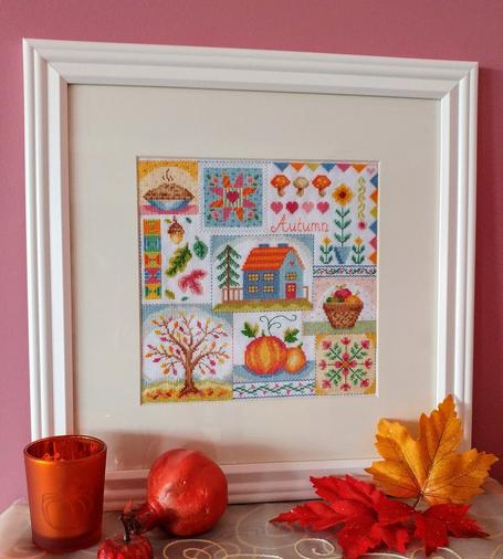 A framed cross stitch piece of several autumn motifs, such as a pie, some pumpkins, a blue house with orange roof, a basket of apples, and little quilt squares.