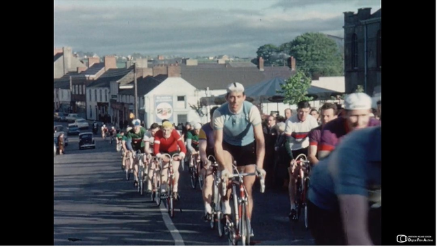 Archive image from 1960. It's in colour and shows a group of cyclists going uphill in a small town in Northern Ireland.