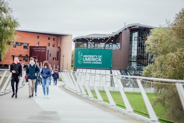 A photograph taken on a white pedestrian bridge being used by several groups of people. In the background there are two large buildings, one with orange bricks and a square wooden box. The other is glass-fronted, with a wooden curved section and a green University of Limerick banner. The bridge is flanked by large mature trees