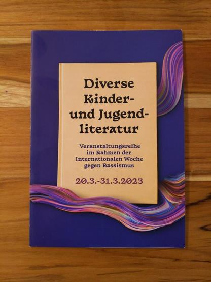 Foto eines Flyers der auf einem Holztisch liegt: "diverse Kunder- und Jugendliteratur" Veranstaltungsreihe im Rahmen der internationalen Wochen gegen Rassismus 20.3.-31.3.2023