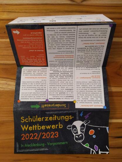 Foto eines Flyers der auf einem Holztisch liegt: "Schülerzeitungs-Wettbewerb 2022/2023 in Mecklenburg-Vorpommern"