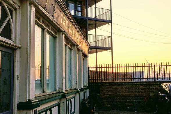 A worn pub frontage reads 'The Old Sun', the letters reflecting against the setting sun - there's further text but it's too indistinct to be read. The opaque glass front door of the pub says 'Old Sunb' with a picture of a sun between the two words, and a fancy border around the edge of the glass. Beyond the frontage there's a wall and a fence with balconies raising above, and in the far distance cranes and a wind turbine.