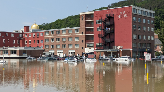 Back of a hotel, with a number of cars half submerged in a flooded parking lot. A few people are standing on the fire escape. In the background, a gold dome and green forested hill.