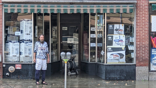 A man in a tie-dyed shirt smokes a cigarette in front of a hardware store. On the bricks and cardboard boxed in the window you can see a high water line, 3-4 feet above the sidewalk.