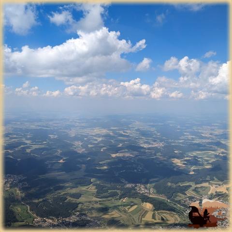 Foto aus dem Flugzeug heraus auf die Landschaft ca 1400m tiefer, darüber einige Wolken am Himmel.