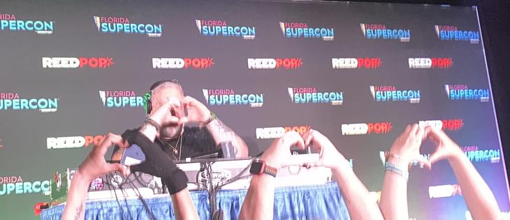 Kristian Nairn sitting behind his DJ set-up doing heart-hands facing out to the crowd, and three sets of people facing him doing heart-hands back at him