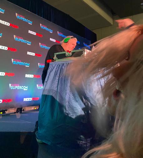 Picture of Kristian Nairn from the side at his DJ set up and a blurry shot of a blonde woman dancing in the foreground