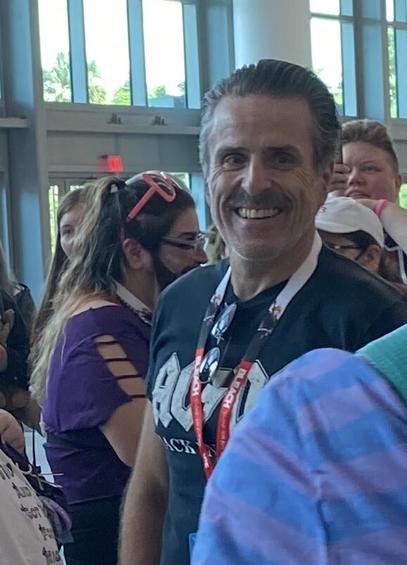 A picture of a smiling Con O'Neill in a black AC/DC tshirt in front of a small crowd of people in the background