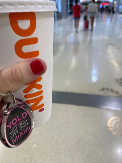 A hand with a red fingernail holding a large Dunkin Donuts coffee cup and a keychain that says "YOLO You only live once Miami" taken in an airport