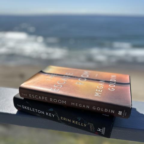 Two books sitting on a rail overlooking the ocean. They are “The Escape Room” by Megan Goldin and “The Skeleton Key” by Erin Kelly
