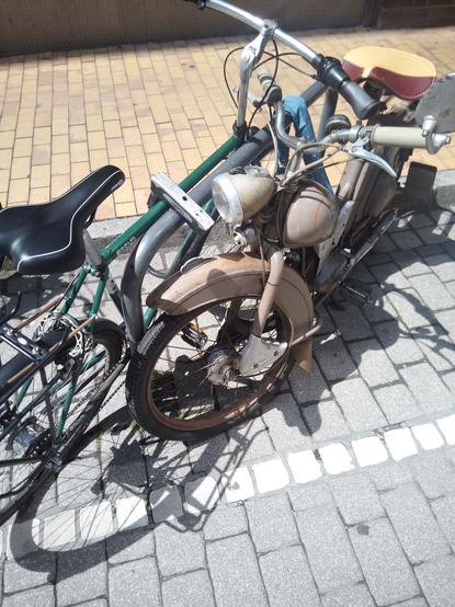 Ein Radbügel in der Stadt. Links steht ein grünes Herrenrad. Rechts ein sehr altes DDR-Moped der Marke Simson aus Suhl in Thüringen.
