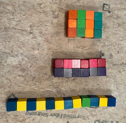 Wooden colored colored cubes showing 3 ways to factor the number 12. 
Top: green and orange arranged in 3 by 4 array (2 columns of 3 orange cubes alternating with columns of 3 green cubes
Middle: 6 pink cubes in a horizontal row above 6 purple cubes, making 2 x 6
Bottom: 12 cubes in a line, alternating 7 dark colors with 5 yellow cubes.