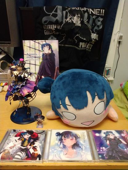 A collection of Yoshiko Tsushima/Yohane merchandise. In front are her three solo albums. Behind them are a figure, a small acrylic stand, and a nesoberi. Behind those are a copy of Aqours magazine with Yohane on the cover, and a bag with a picture of her on the front.