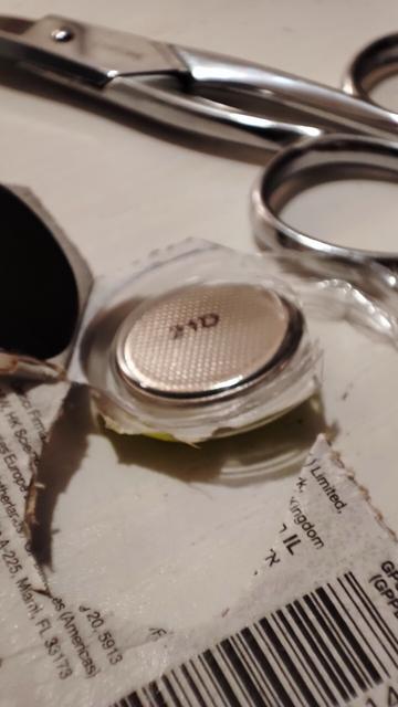 A CR2032 battery inside the transparent casing that is partially cut. In the background, a pair of scissors.