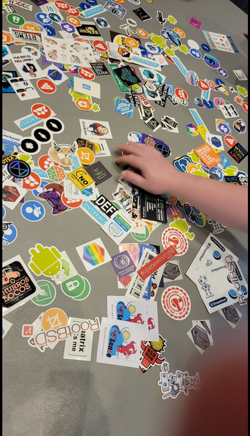 A table covered in stickers. In the bottom right corner you can spot a few BSD, Perl, OSMI, and even Dreamhost stickers.
