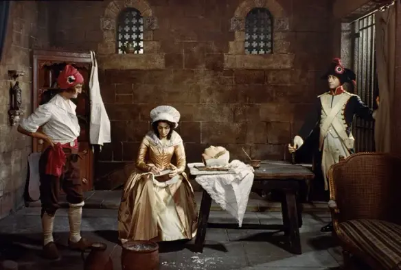 Photograph of a display in Madame Tussauds in London where a young Marie is mixing her paste while the decapitated head lays on the table next to her. A Revolutionary soldier stands on the right at the door and another man is standing to her left with her hands on his hips.