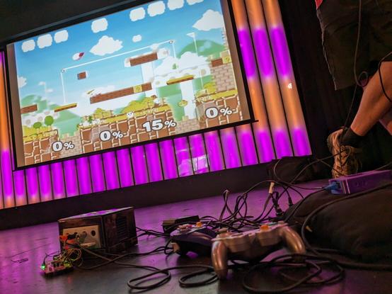 Stage of SummerCon at Littlefield NYC with a modified GameCube playing Super Mash Bros Melee, circuits and wires hanging out.