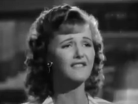 B&W film frame of a  blonde-haired woman with a painfully sad face and tears has her upper teeth on her lower lip (A labiodental fricative, which forms the sounds for the letters “V” and “F”.)