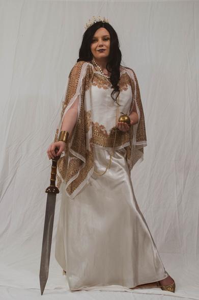 A white woman in a white and gold chiton-like costume poses, leaning on a sword, and holding an golden apple. A pearly crown graces her head and strands of prarls hang from her neck.