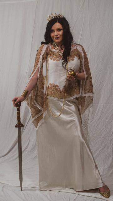 A white woman in a white and gold chiton-like costume poses, leaning on a sword, and holding an golden apple. A pearly crown graces her head and strands of prarls hang from her neck.
