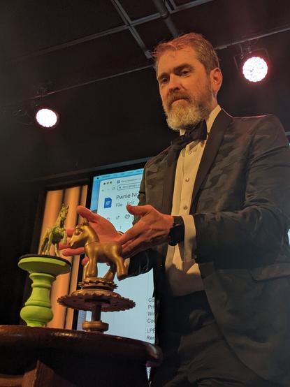Host at SummerCon on stage in a suit and tie presenting the actual physical Pwnie Awards on a table.