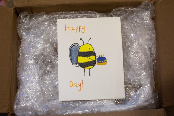 Card with painted bee holding a birthday cake, situated in between the words, "happy", and "day". I.e., "Happy Bee Day".