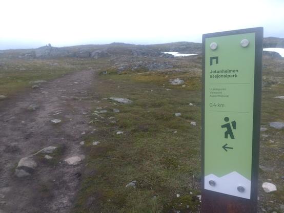 Til venstre en sti som går oppover bakke. Til høyre et grønt skilt med teksten "Jotunheimen nasjonalpark. Utsiktspunkt. Viewpoint. Aussichtpunkt. 0.4km"