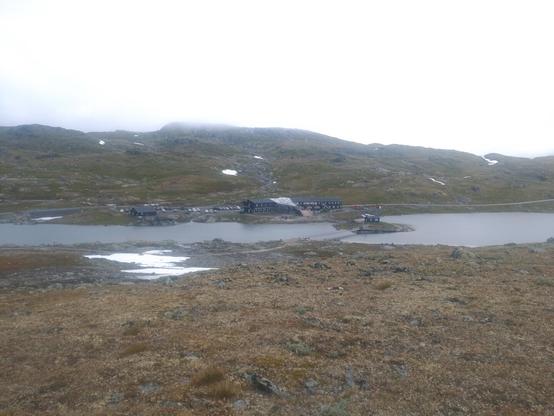 I midten et vann med en holme. Mellom holmen og fast land går en langt smal gangbru. I bakgrunnen er to mørke toetasjes trebygninger knyttet sammen med en glasskuppel. Bak bygningen går en asfaltert vei. Bak veien ligger lave runde fjell.