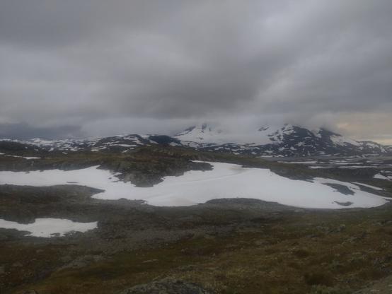Høye og spisse fjell strekker seg opp fra flatere omgivelser med toppene dekket av lave skyer og sidene dekket av isbreer.