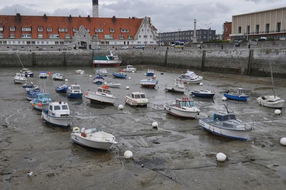 Das Hafenbecken von Calais, 
trockengefallen bei Ebbe.
Im Hafenschlick liegen zahlreiche kleine Fischkutter sowie private Segelboote und Yachten.
Im Hintergrund ein historischer Hafenspeicher mit einem hohen Ziegeldach, vielen Dacherkern und Renaissancegiebel