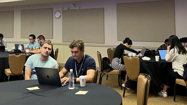 A conference room with people sitting around tables and working on their laptops
