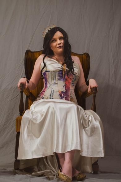 A white woman in a silver dress and corset wearing a pearly crown and a necklace of golden chains. She sits on a golden throne against a white backdrop.