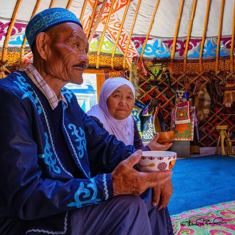 A Mongolian couple wearing colorful traditional clothing share soup with a group of travellers in an elaborate ger