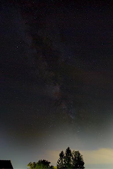 Photograph of a dark starry night sky with the milky way visible in the middle, the sky getting lighter towards the bottom. There are the tops of two trees and the edge of a roof visible at the lower edge and a cloud that is passing behind the trees.