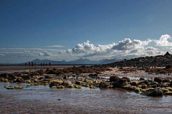 2016: Eine Gruppe von Menschen geht an einem Strand spazieren. Im Vordergrund sind Wasser und bemooste Steine zu sehen, das Wasser glitzert durch den Sonnenschein. Im Hintergrund sind Berge zu erkennen. Der Himmel ist blau mit einigen weißen Wolken.

2016: A group of people are walking on a beach. In the foreground water and mossy stones can be seen, the water glistens through the sunshine. Mountains can be seen in the background. The sky is blue with some white clouds.