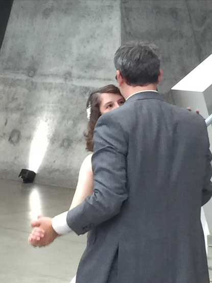 Color image of a bride and groom having their first dance. Couple is centered, holding hands, bride's eyes on her new husband. The modern architecture of a modern art museum, in concrete, granite, and stainless steel sets the background, with a single spotlight.