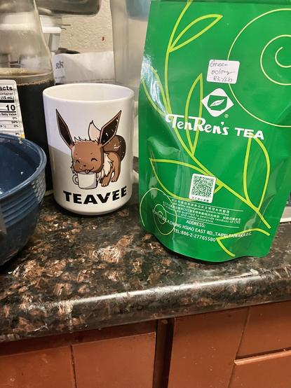 a white mug with a picture of a brown dog like creature with ears like a rabbit.  below that is the word TeaVee.  Next to it is a green bag of tea with yellow swirls and leaves with the logo of tenren’s Teas