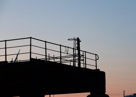 A short ladder with a hoop at the top is mounted against a pole by the safety rails on a raised platform. There is a sunset sky behind, red towards the bottom, blue near the top.