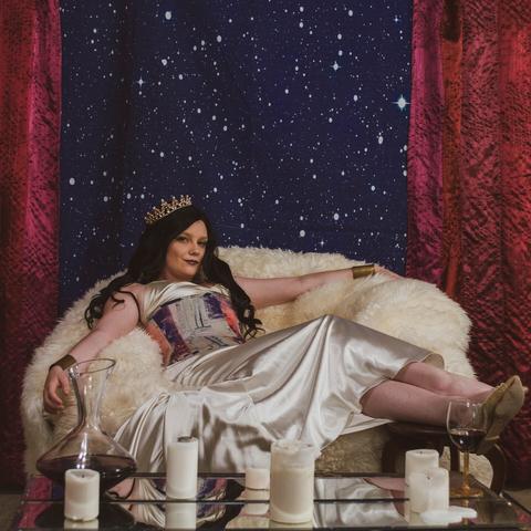 A white woman with long dark hair sits on a fur-covered throne against a backdrop of stars. She wears a pearly crown and a colorful corset over a silver dress. In the foreground sit candles and a decanter of red wine.