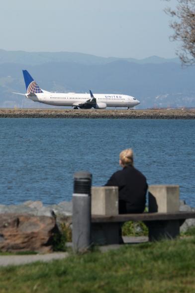 Telephoto shot of a woman watching an airplane across a body of water.