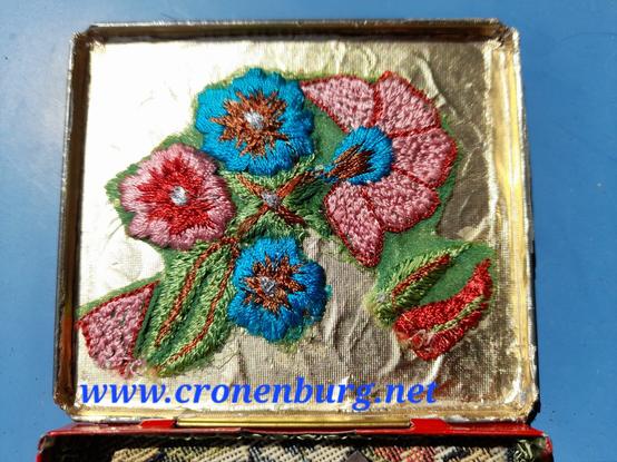 The inside of the lid is lined with the original golden cigarette paper. A bouquet of embroidered flowers is fashion waste from saree production. It is colourful and machine-stitched.